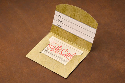 An open gift card holder, resting on a brown surface, showcases the Shaving Class Gift Card inside. The holder features spaces labeled To, From, and Value—making it an ideal gift for anyone interested in straight razor shaving or learning the traditional art of using a safety razor.