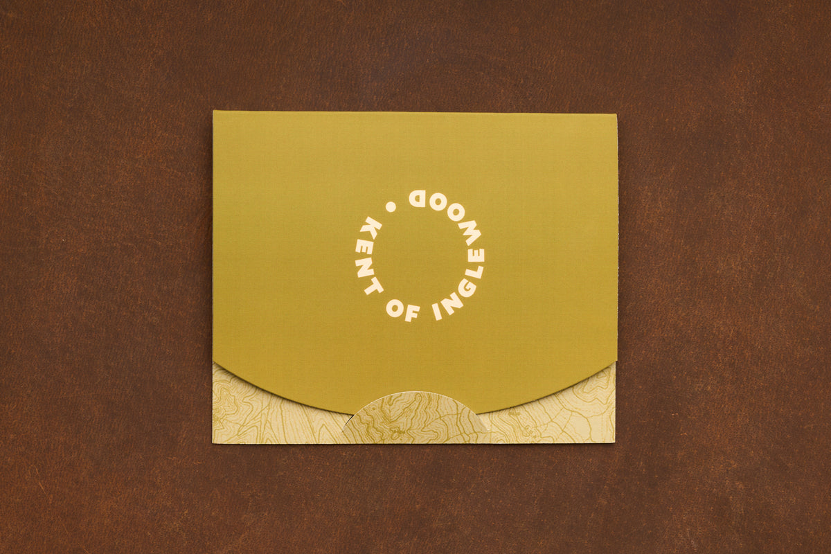 A green and tan envelope with “KENT OF INGLEWOOD” written in a circle on the flap, designed for presenting the Shaving Class Gift Card or introducing someone to straight razor shaving, placed on a brown textured surface.