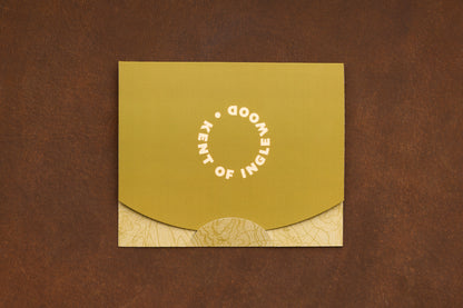 A green and tan envelope with “KENT OF INGLEWOOD” written in a circle on the flap, designed for presenting the Shaving Class Gift Card or introducing someone to straight razor shaving, placed on a brown textured surface.