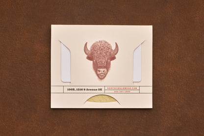 A cream envelope featuring a buffalo illustration, business address, and contact details rests on a brown surface. The envelope displays two flaps and a partial gold seal at the bottom—an ideal presentation for the Shaving Class Gift Card or other grooming essentials like straight razor shaving tips.