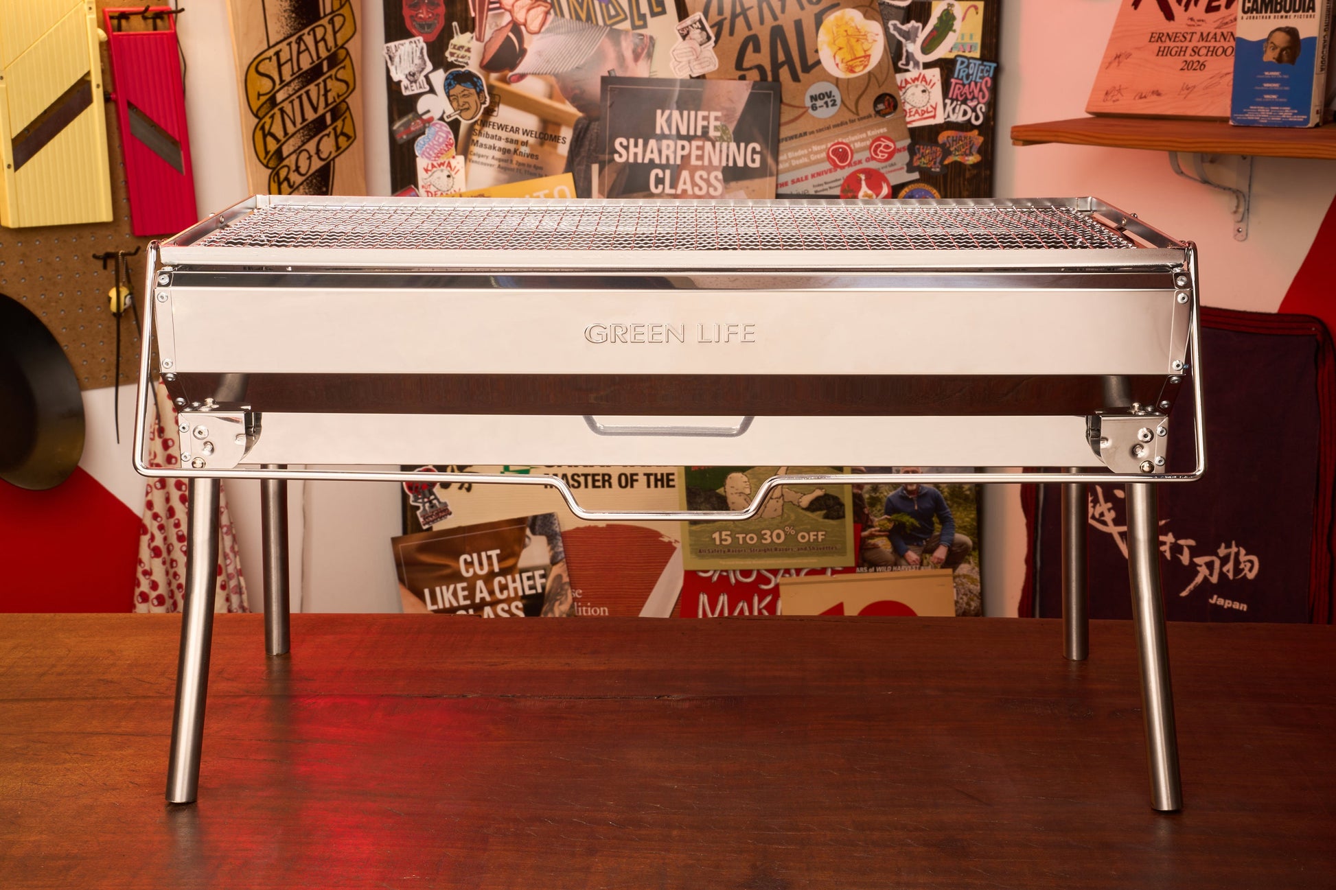 A Green Life Stainless Steel BBQ Grill stands on a wooden surface, its sleek design complemented by colorful posters and kitchen tools in the background—ideal for anyone who enjoys stylish charcoal grilling.