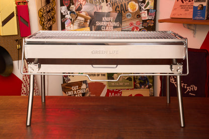 A Green Life Stainless Steel BBQ Grill stands on a wooden surface, its sleek design complemented by colorful posters and kitchen tools in the background—ideal for anyone who enjoys stylish charcoal grilling.