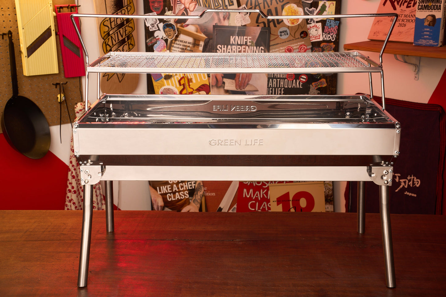 A Green Life Stainless Steel BBQ Grill, with its compact design reminiscent of a portable konro grill, stands on a wooden table in front of a wall adorned with posters and kitchen tools. Its mesh rack is ideal for charcoal grilling, combining style and versatility as a dependable Stainless Steel BBQ Grill for any kitchen or outdoor setting.