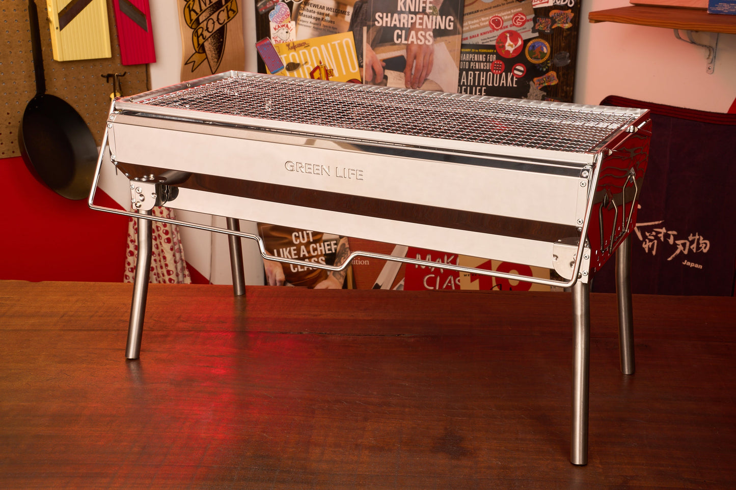 A shiny, rectangular Green Life Stainless Steel BBQ Grill stands on a wooden table in a room decorated with various culinary posters and cooking tools.