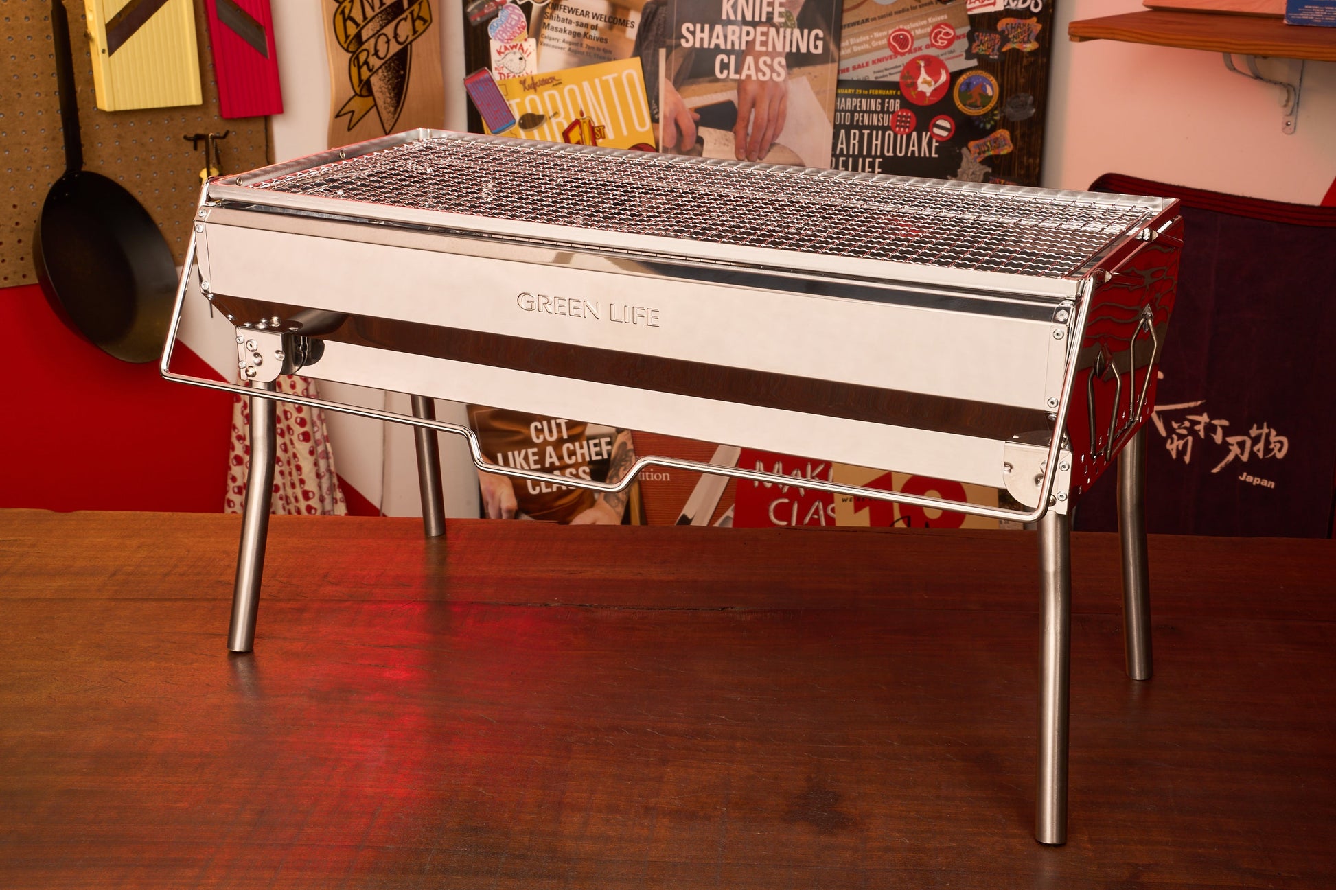 A shiny, rectangular Green Life Stainless Steel BBQ Grill stands on a wooden table in a room decorated with various culinary posters and cooking tools.