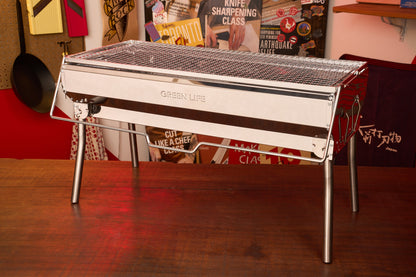 A shiny, rectangular Green Life Stainless Steel BBQ Grill stands on a wooden table in a room decorated with various culinary posters and cooking tools.