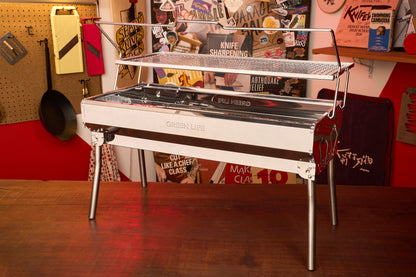 A Green Life Stainless Steel BBQ Grill stands on a wooden table in a colorful room decorated with posters, tools, and kitchen items on the walls.