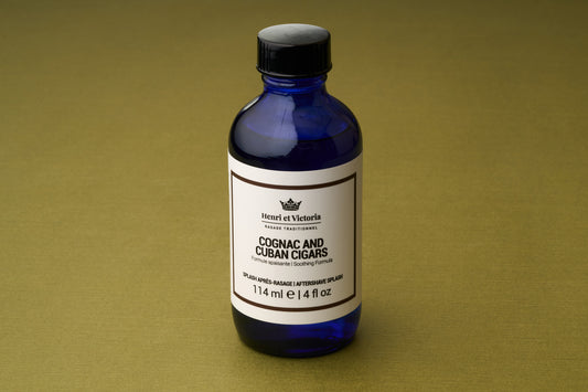 A blue bottle labeled “Henri et Victoria - Cognac and Cuban Cigars Aftershave Splash,” 114 ml, rests on a simple gold surface.