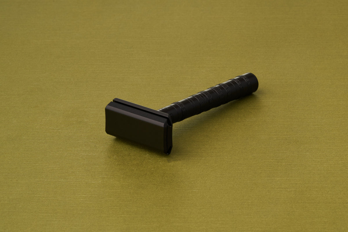 A Henson Safety Razor Aluminum Jet Black with a textured handle rests on a flat olive-green surface.