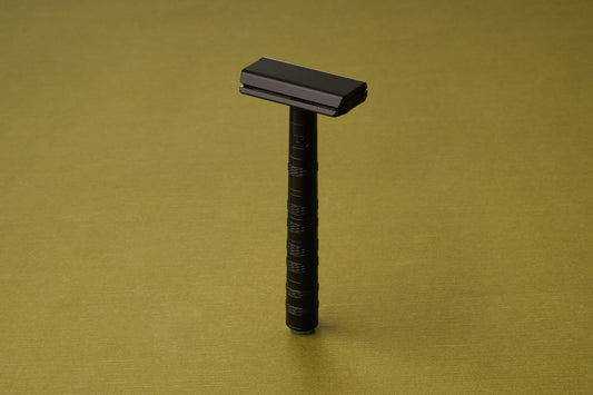 The Henson Safety Razor Aluminum Jet Black stands upright on a textured olive-green surface, its sleek black frame casting a subtle shadow.