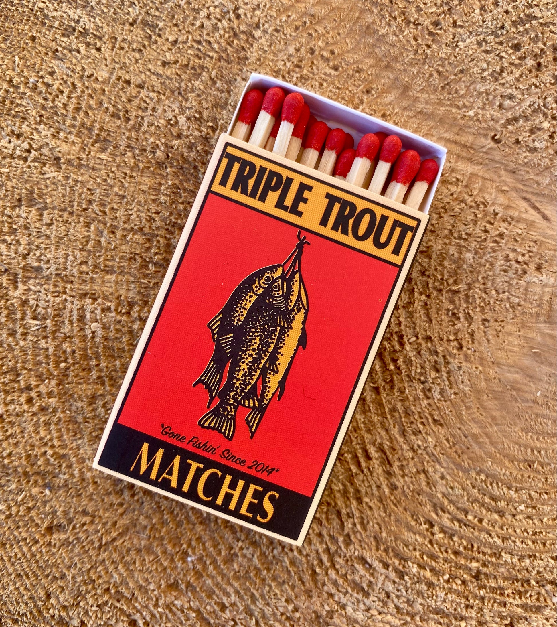 Matchbox labeled 'Triple Trout Matches' with red matches on a wooden surface