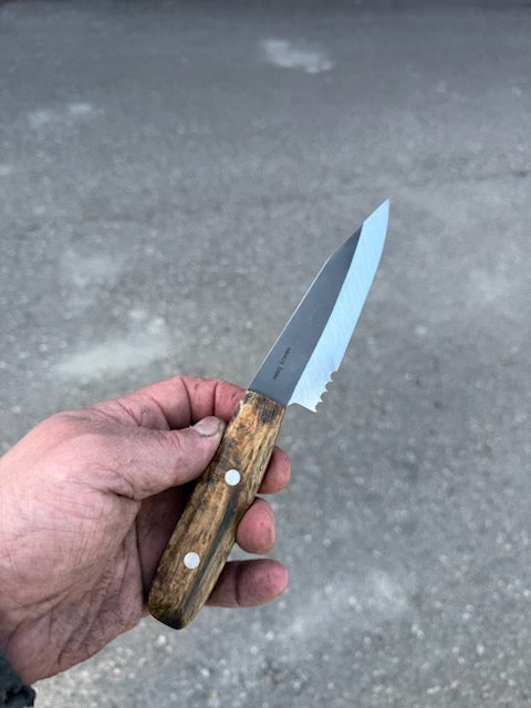 A hand holding the RESQ Knives Korpi Camp Kitchen Knife, featuring a wooden handle and shiny recycled stainless steel blade, with a blurred outdoor pavement background.