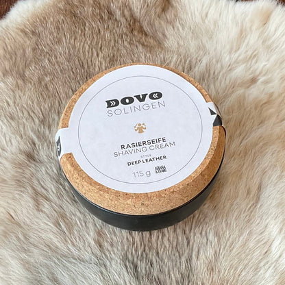 Dovo Deep Leather Shaving Soap in a cork-lidded tin on a fur-like surface, featuring handmade craftsmanship with Oud, Mimosa, and Leather Suede notes.