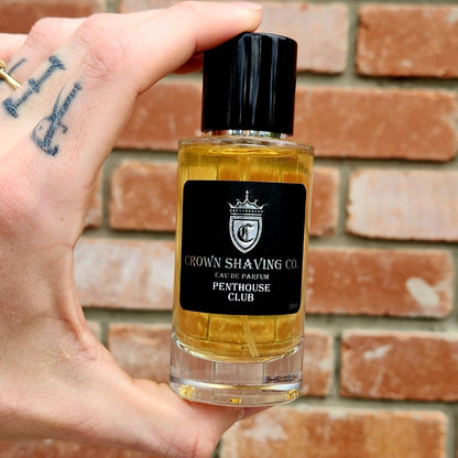 Hand holding Crown Shaving Co. Penthouse Club Eau de Parfum 50mL, clear glass bottle with black label and cap, showcasing golden liquid.