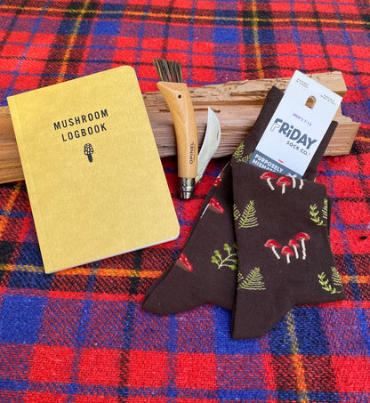 Mushroom Hunter Bundle featuring a yellow logbook, Opinel mushroom knife, driftwood, and Friday Sock Co. socks with red mushrooms and green ferns on a tartan blanket.