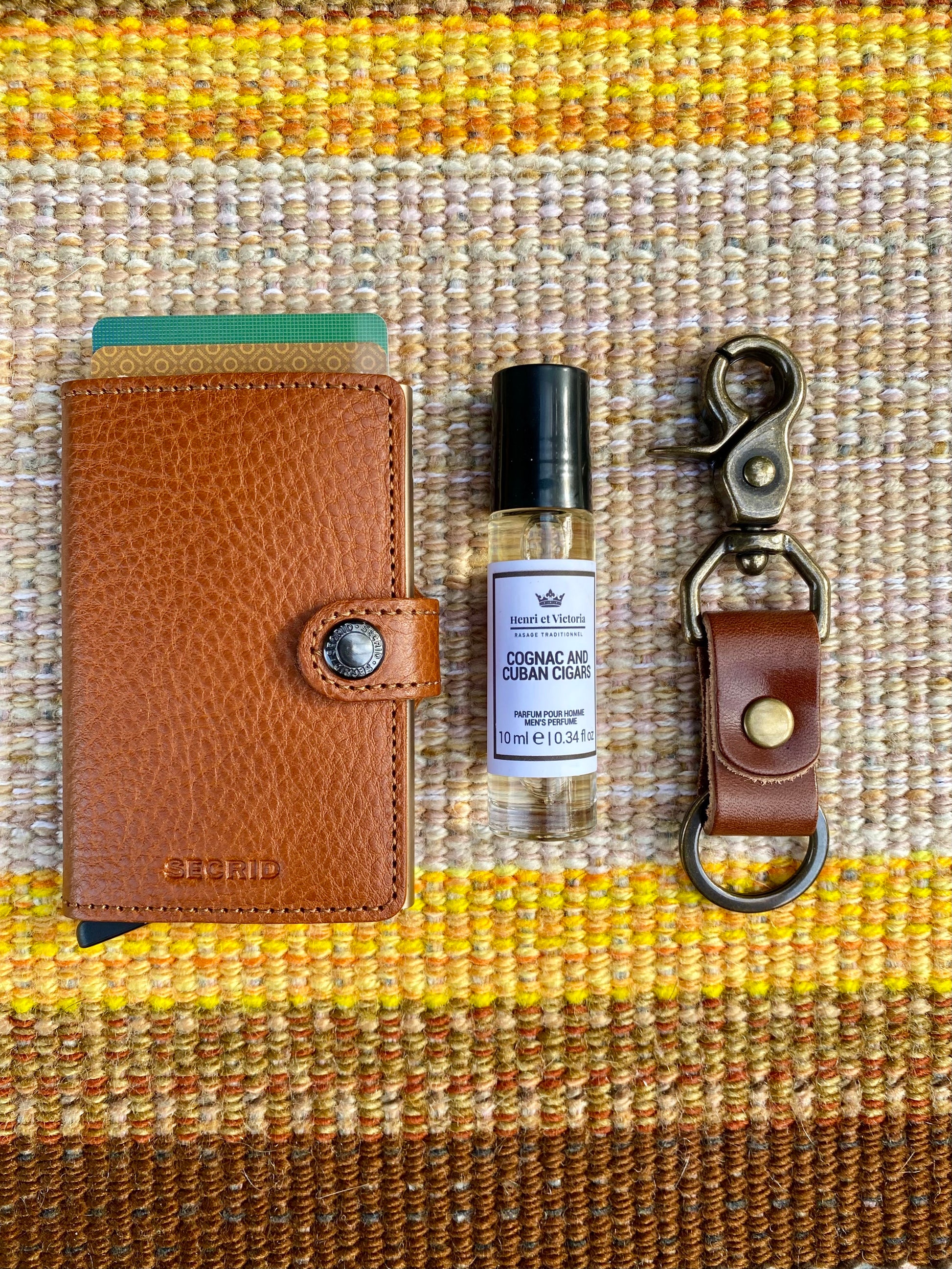 Brown leather wallet, small bottle of cologne, and keychain on a textured fabric background