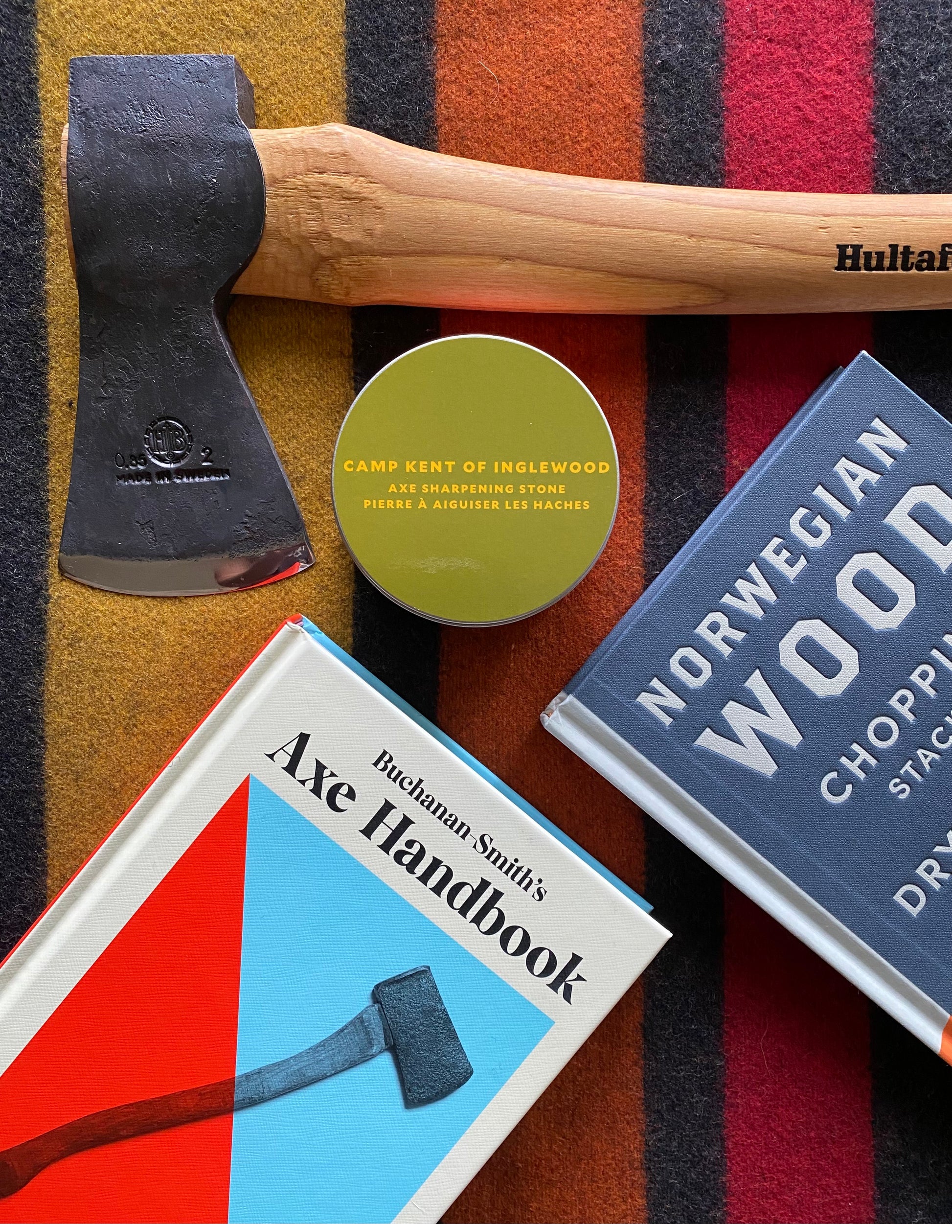 Axe, a book titled 'Axe Handbook', a can of Camp Kent of Inglewood, and a book titled 'Norwegian Wood' on a striped background.