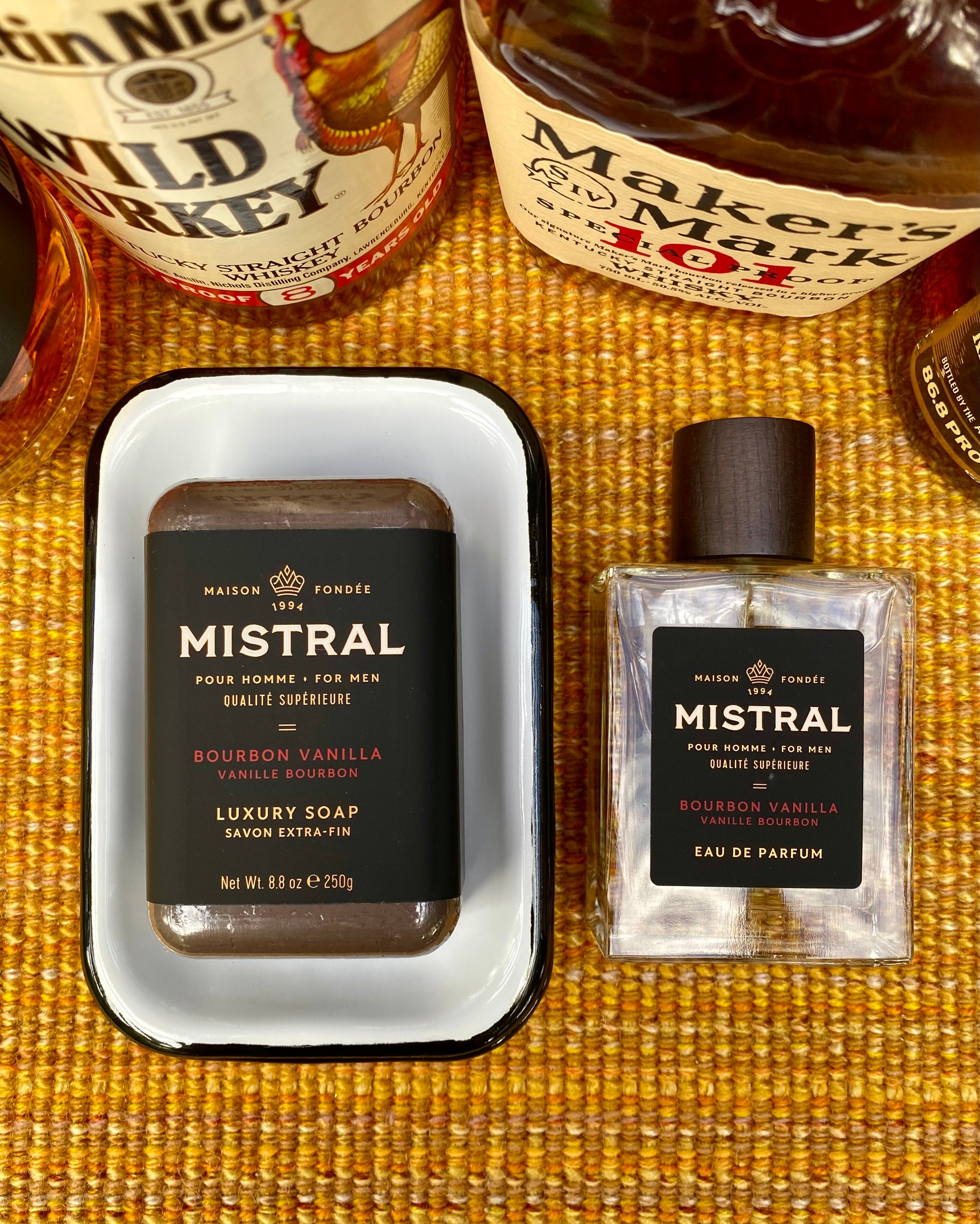 Mistral bourbon vanilla soap and eau de parfum on a textured surface with bottles of bourbon in the background.