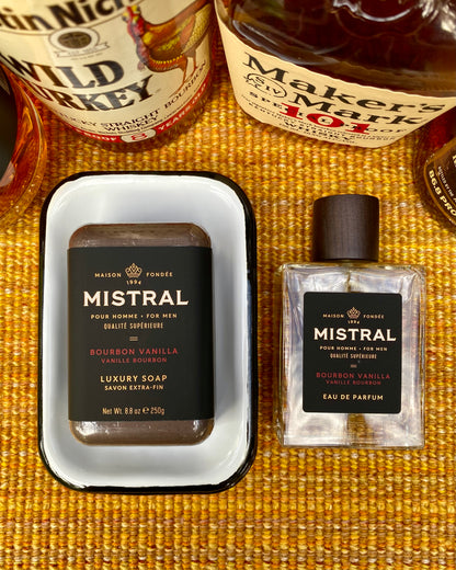 Bourbon Boss Bundle: two bourbon bottles with Mistral Bourbon Vanilla soap in a dish and matching eau de parfum on a mustard-yellow surface.
