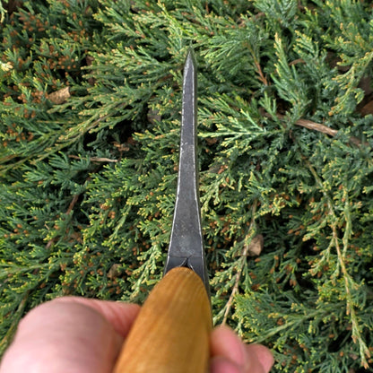 A hand grips the Hultafors Ekelund Small Felling Axe, featuring an American hickory handle and pointed metal head, poised toward dense green foliage.