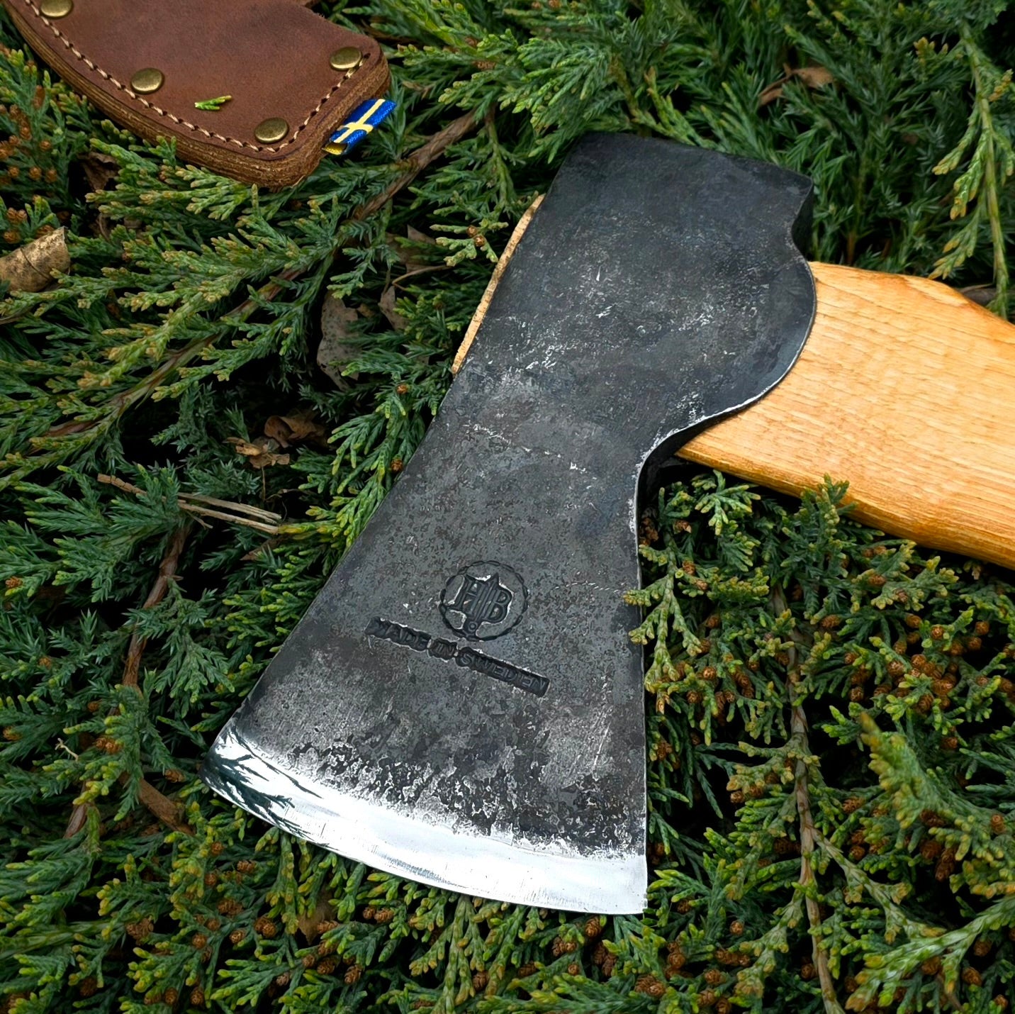 The Hultafors Ekelund Small Felling Axe with an American hickory handle and Swedish carbon steel head, marked with branding, rests on green foliage. A leather sheath with blue and yellow details lies beside the weathered tool.