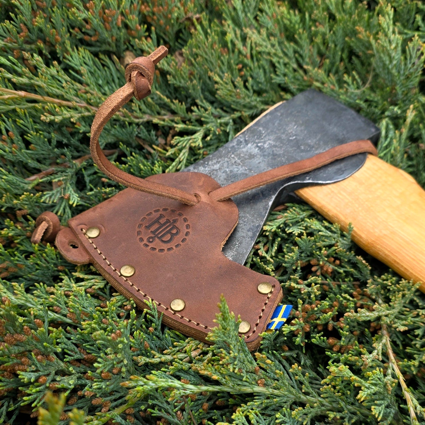 A Hultafors Ekelund Small Felling Axe with an American hickory handle and a brown leather sheath—featuring a stamped logo, brass rivets, and a Swedish flag tag—rests on green juniper branches.