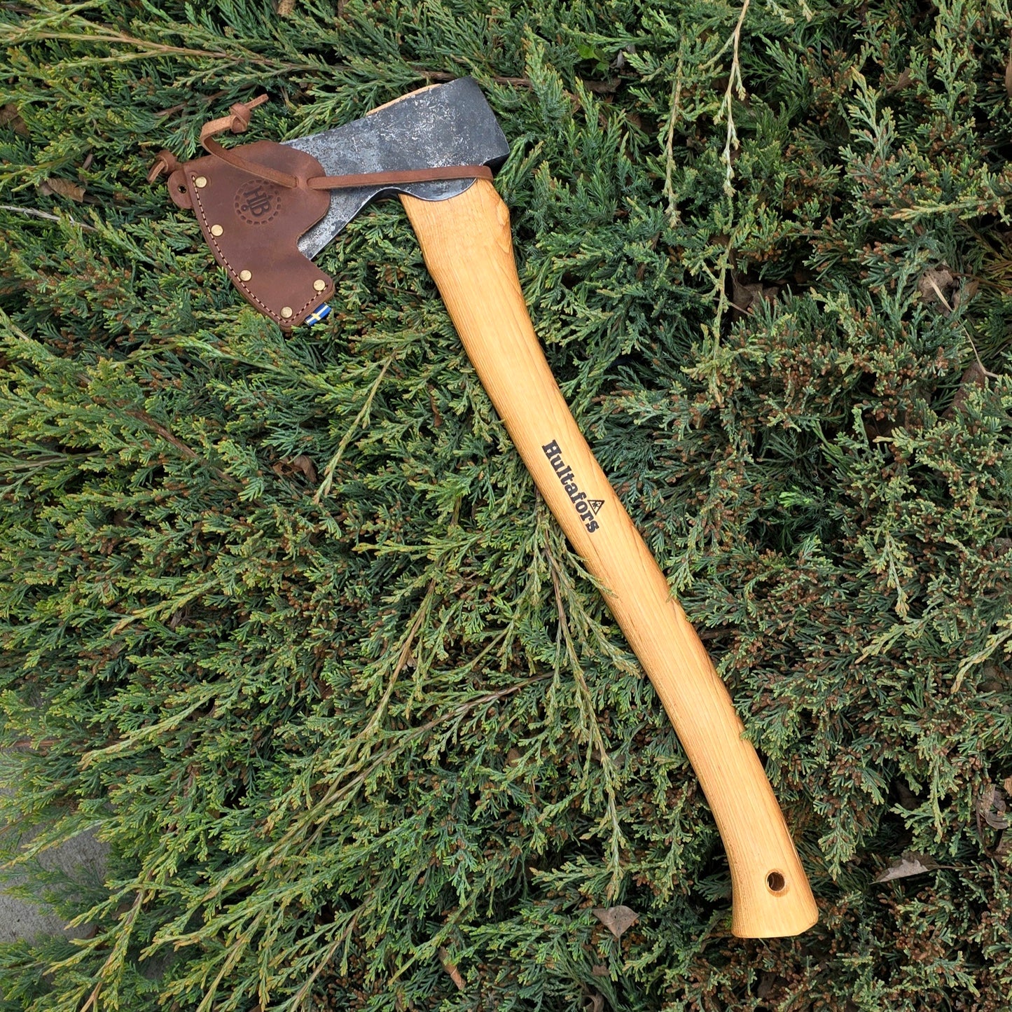 The Hultafors Ekelund Small Felling Axe, featuring a Swedish carbon steel blade and American hickory handle, rests on dense green foliage with its leather sheath covering the edge.
