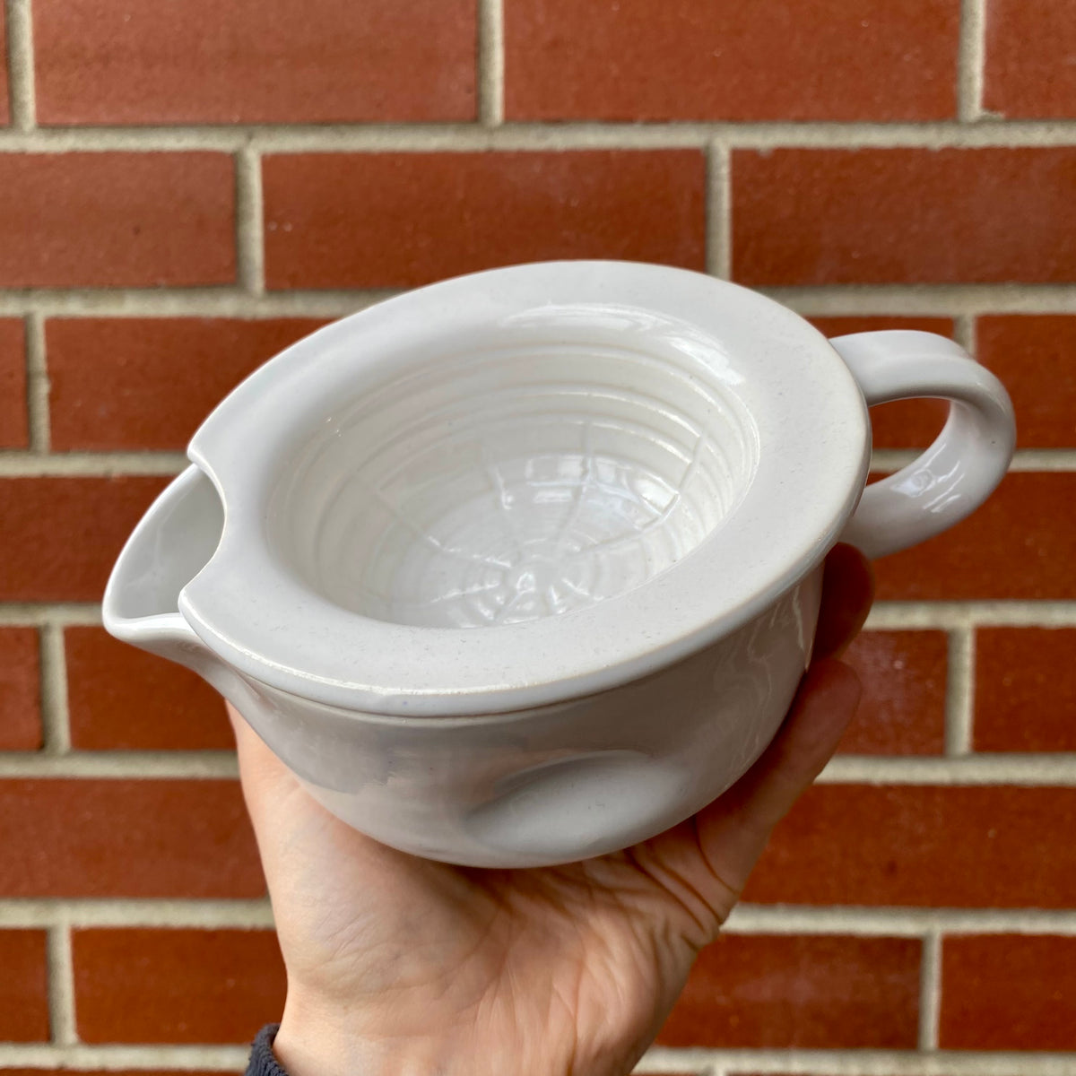 Lot. 15 Scuttle Style Shaving Bowl held in hand, showcasing its ceramic design with handle, pour spout, and interior ridges for enhanced lathering.