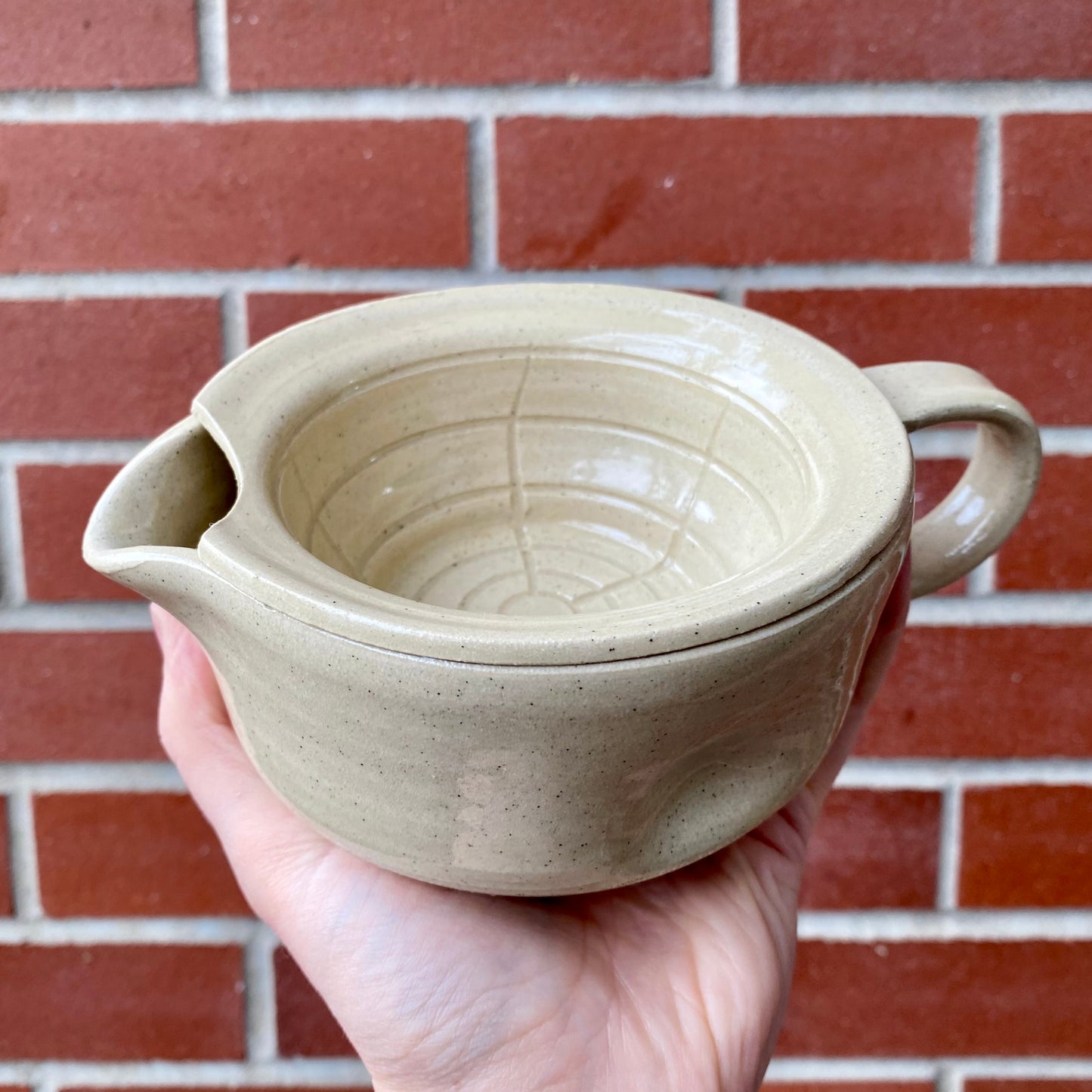 Hand holding a beige ceramic pot with two handles against a brick wall.