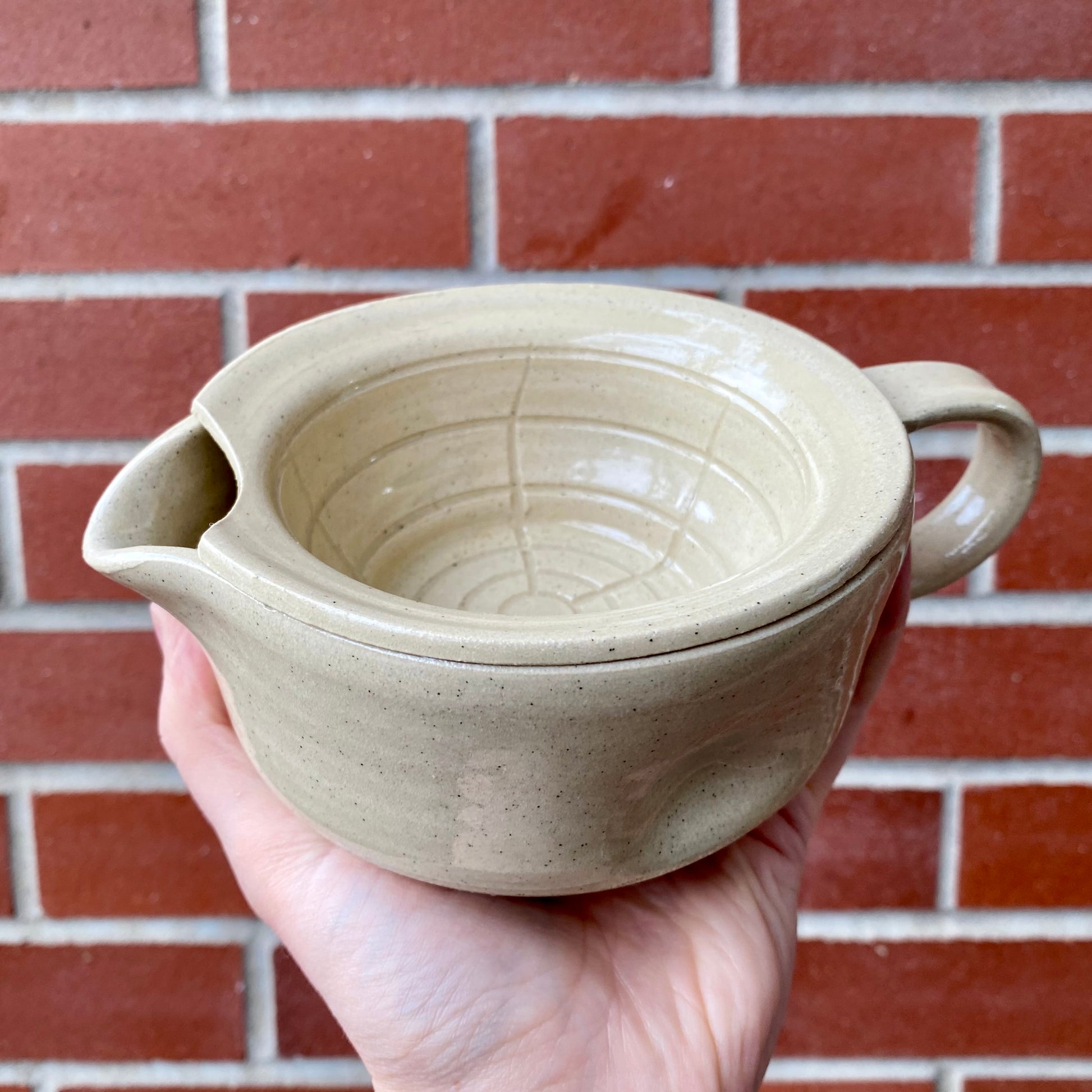 Hand holding a beige ceramic pot with two handles against a brick wall.