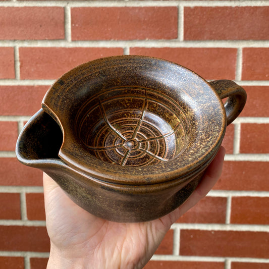 Hand holding a brown ceramic strainer against a brick wall