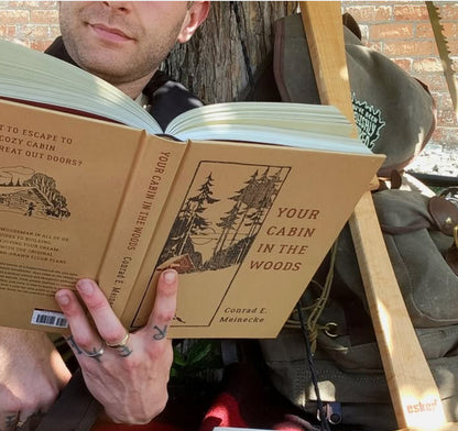 Person reads Your Cabin in the Woods by Conrad E. Meinecke, surrounded by cabin-building tools, embodying a rustic, nature-inspired lifestyle.