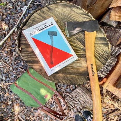 Buchanan-Smith’s Axe Handbook on a tree-stump with a small splitting axe, green canvas apron, wood logs, and sneakers, showcasing tools for outdoor enthusiasts.
