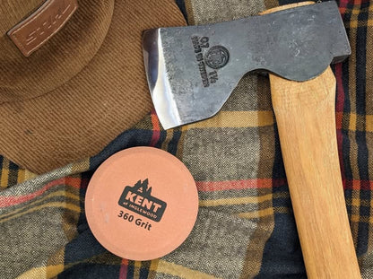 Kent of Inglewood Axe Sharpening Stone on plaid cloth, alongside a corduroy cap and axe, highlighting its two-sided grit for maintaining sharp edges.