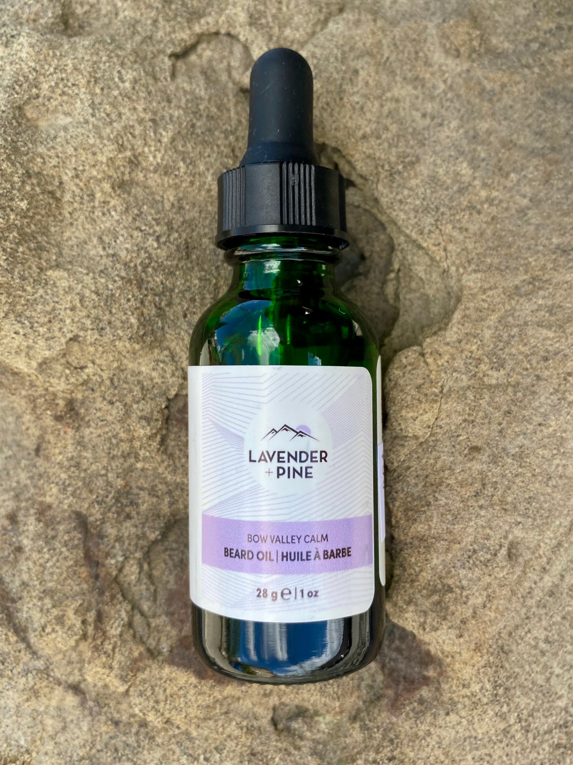 A green glass dropper bottle of Banff Beard Co. Lavender + Pine Beard Oil rests on textured stone, its white label with purple accents showing a size of 28g (1 fl oz).