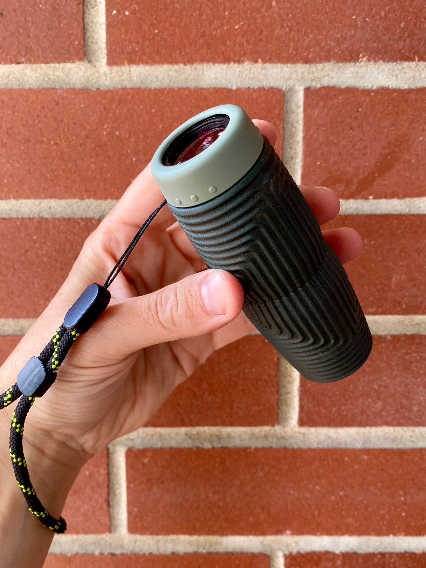 A hand holding the NOCS Provisions Zero Tube 10x25 Monocular—a compact black and green waterproof monocular with a wrist strap—positioned in front of a red brick wall.