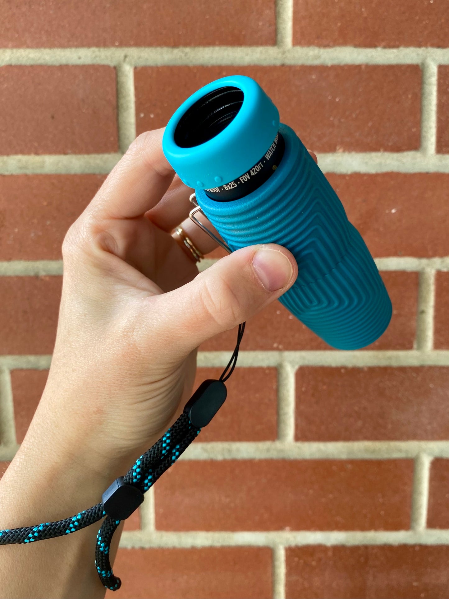 A hand holding a NOCS Provisions Zero Tube 10x25 Monocular in blue, with a black wrist strap, in front of a red brick wall.