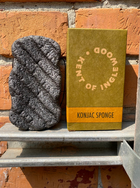 A black Kent of Inglewood Konjac Body Sponge, displayed with its green and orange box labeled “Konjac Sponge,” sits on a metal shelf against a brick wall in bright sunlight, providing a biodegradable bath sponge alternative for a plastic-free shower.