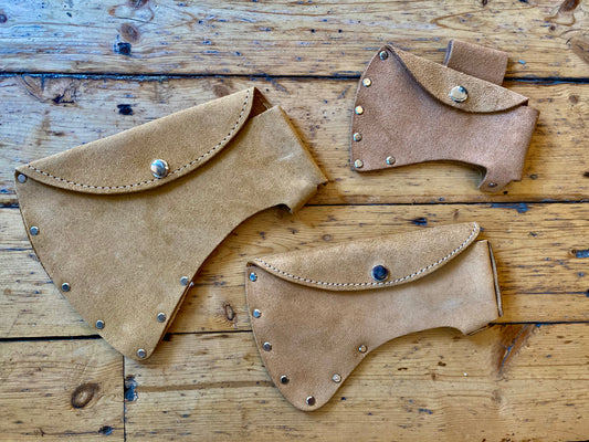 Three G. Hjukstrom Leather Axe Sheaths made from tan split leather with metal rivets and snap buttons are displayed on a rustic wooden surface, each featuring a sturdy belt loop design for secure and convenient carrying.