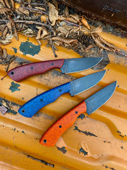 Three Rainy Day Forge Cisco small fixed-blade knives, each featuring vibrant red, blue, and orange speckled handles, are arranged side by side on a rusty yellow metal surface scattered with dried leaves. Crafted from carbon steel, these bushcraft knives make an excellent addition to any everyday carry knife collection.