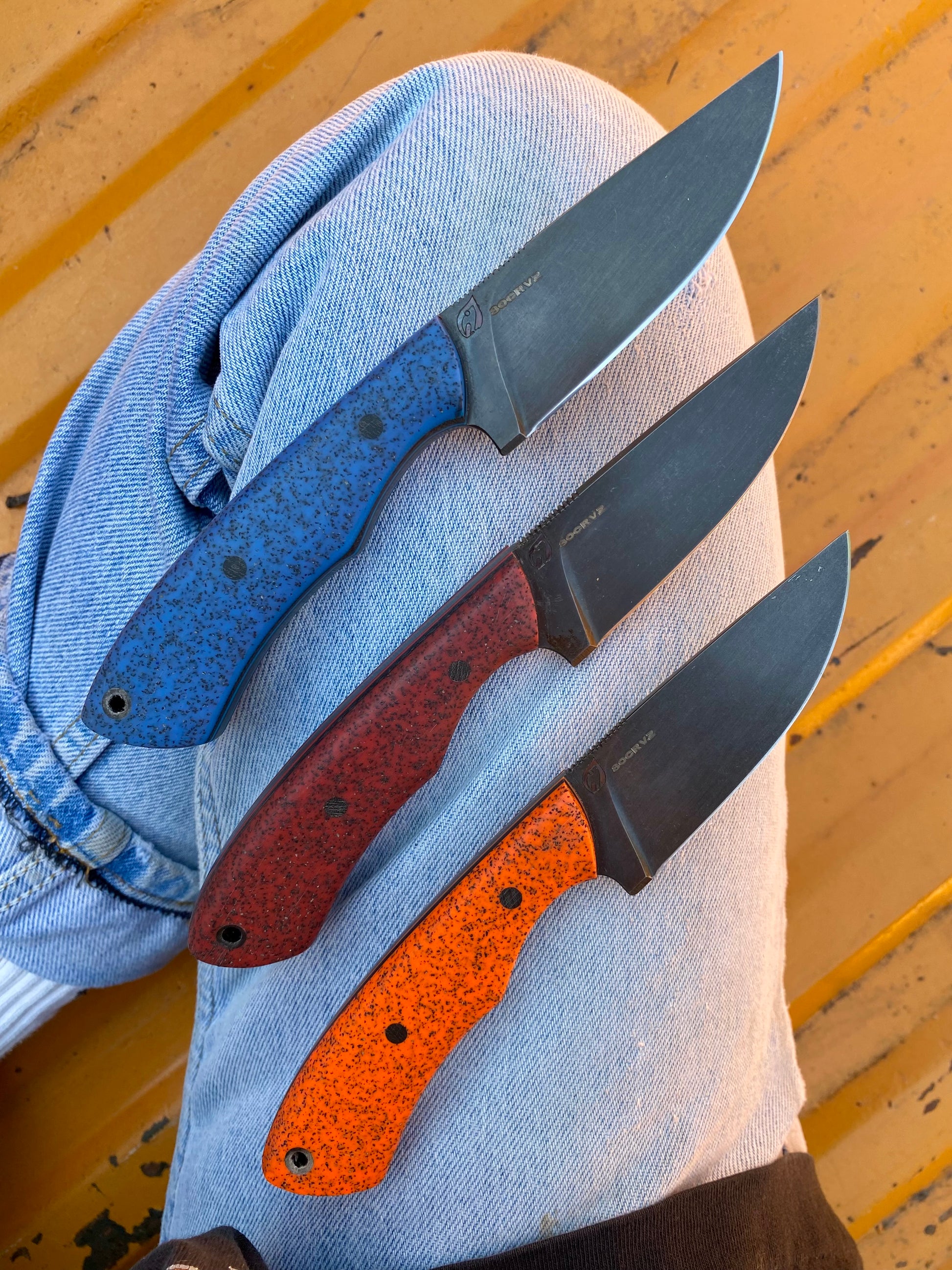 Three fixed-blade Rainy Day Forge Cisco knives—each an everyday carry knife—feature blue, red, and orange speckled handles resting on denim-clad legs against a vibrant yellow background.