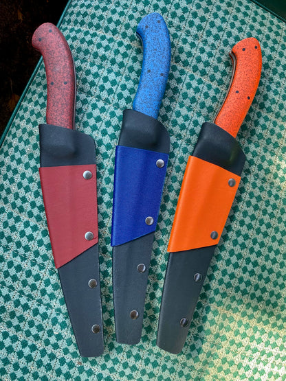 Three Rainy Day Forge Lake Winnipeg Fillet Knives with curved handles in red, blue, and orange are displayed on a green checkered surface. Each knife rests in a matching-colored sheath, capturing the spirit of the Canadian Wilderness.