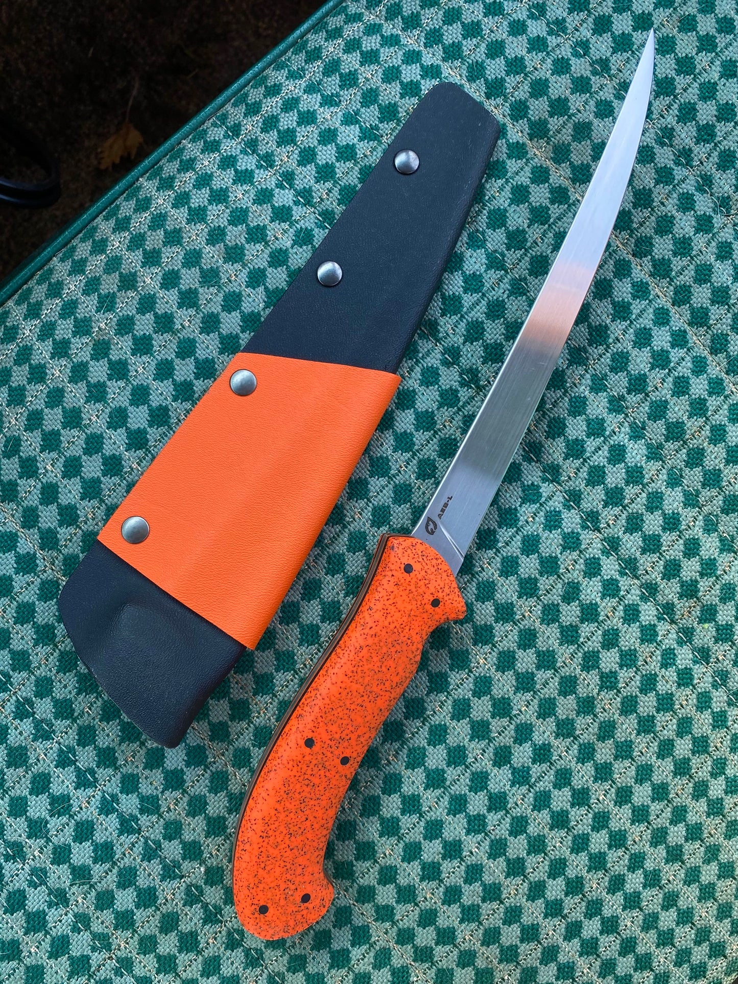 A Rainy Day Forge Lake Winnipeg Fillet Knife with an orange speckled handle rests beside its black and orange sheath on a green checkered fabric surface, evoking the spirit of the Canadian Wilderness.