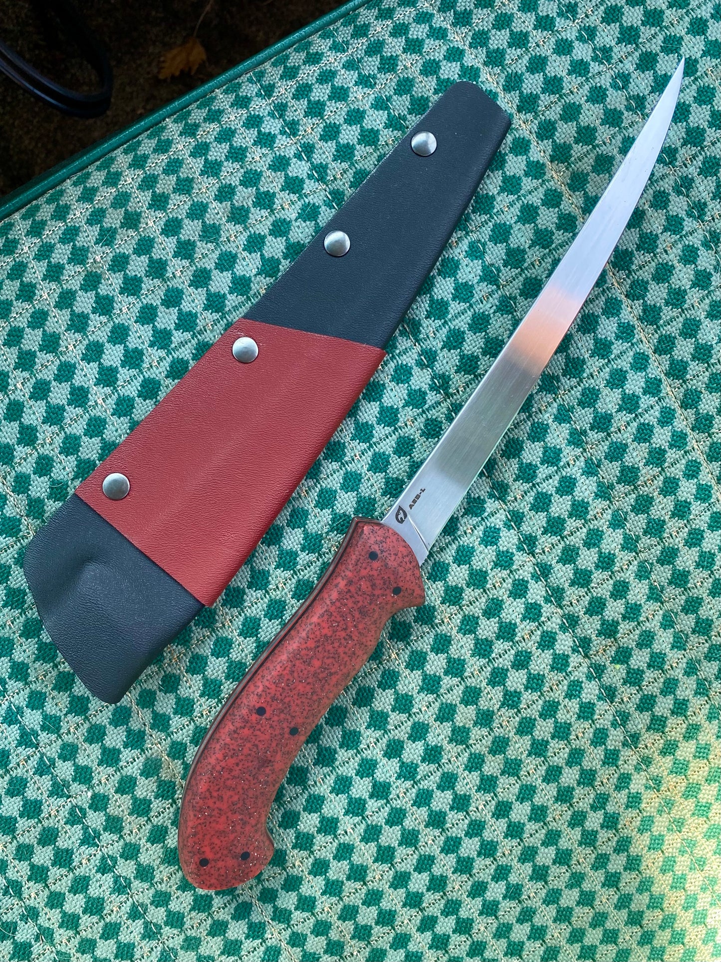 A Rainy Day Forge Lake Winnipeg Fillet Knife, featuring a red speckled handle and a long, thin blade, rests beside its red and black sheath on a green and white checkered surface, capturing the rugged spirit of the Canadian Wilderness.
