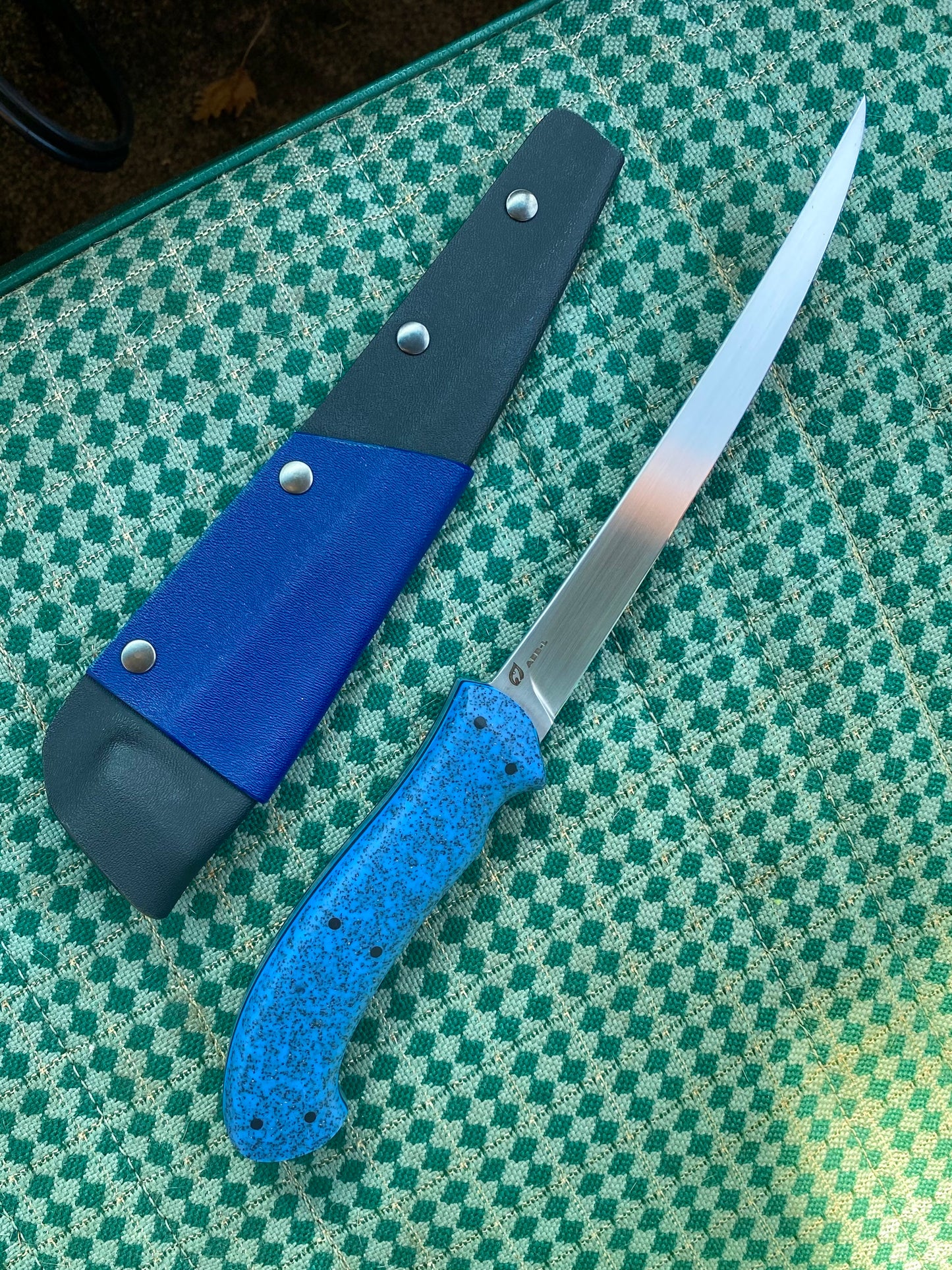 The Rainy Day Forge Lake Winnipeg Fillet Knife, featuring a blue speckled handle and a long, slender blade, rests beside its blue and black sheath on a green patterned surface—perfectly prepared for your next Canadian wilderness adventure.