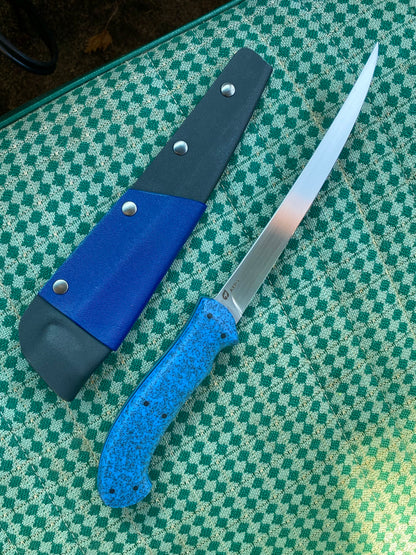 The Rainy Day Forge Lake Winnipeg Fillet Knife, featuring a blue speckled handle and a long, slender blade, rests beside its blue and black sheath on a green patterned surface—perfectly prepared for your next Canadian wilderness adventure.