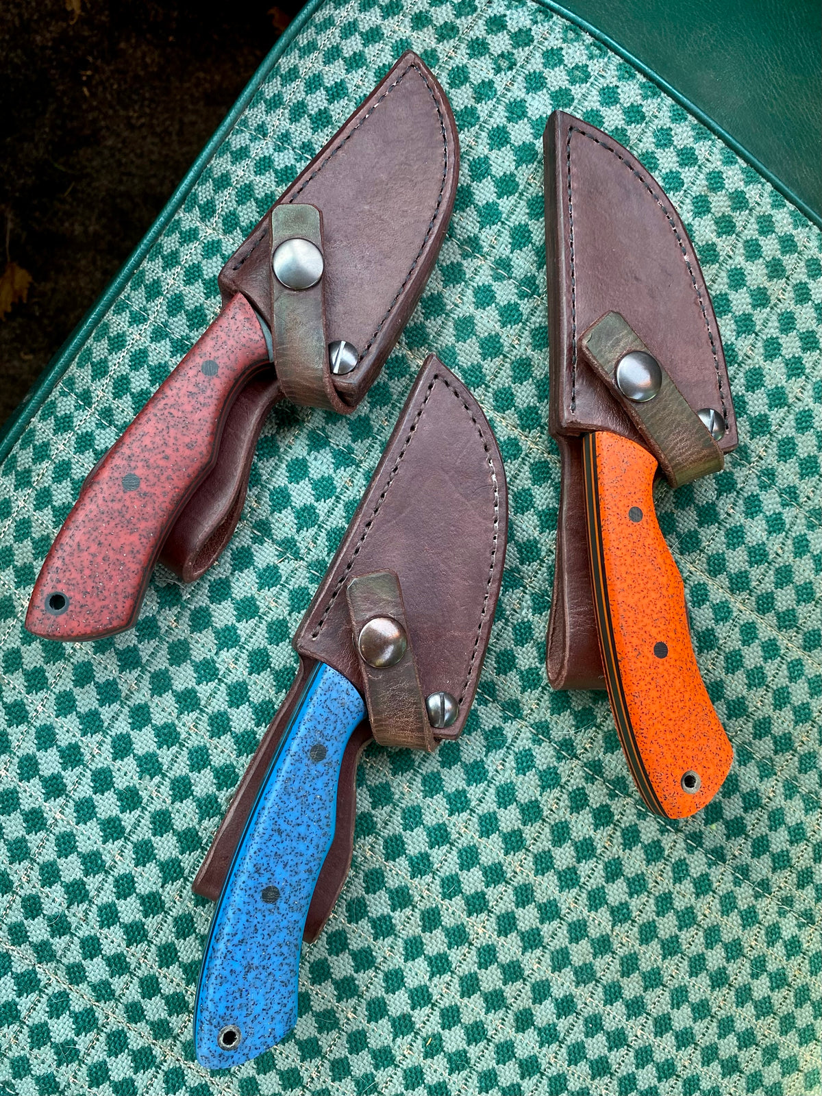 Three knives with red, blue, and orange speckled handles sit in brown leather sheaths on a green and white checkered surface—each handcrafted Rainy Day Forge Cisco carbon steel bushcraft knife, designed for your everyday carry needs.