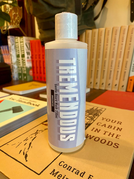 A bottle of Tremendous Beard Wash (250ml), perfect for natural beard care and enhanced with cedar oil, sits on a stack of books—including one titled Your Cabin in the Woods—amid neatly arranged books in the background.