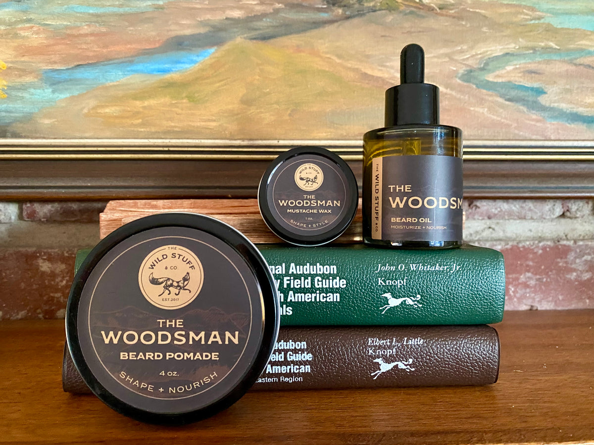 A jar of beard pomade, a tin of mustache wax, and a dropper bottle of Wild Stuff & Co. Woodsman Beard Oil arranged on top of two stacked field guide books.
