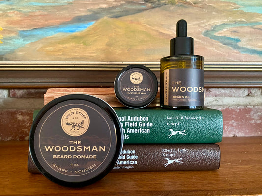 A jar of beard pomade, a tin of mustache wax, and a dropper bottle of Wild Stuff & Co. Woodsman Beard Oil arranged on top of two stacked field guide books.